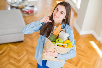 Poster - Beautiful young woman holding paper bag full of healthy groceries surprised with an idea or question pointing finger with happy face, number one