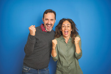 Wall Mural - Beautiful middle age couple together standing over isolated blue background celebrating surprised and amazed for success with arms raised and open eyes. Winner concept.