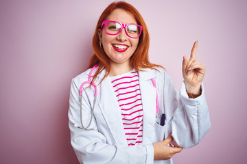 Sticker - Young redhead doctor woman using stethoscope standing over isolated pink background with a big smile on face, pointing with hand and finger to the side looking at the camera.