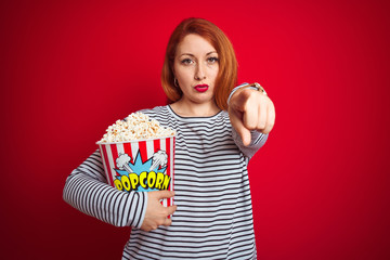 Sticker - Young beautiful redhead woman eating popcorn over red isolated background pointing with finger to the camera and to you, hand sign, positive and confident gesture from the front