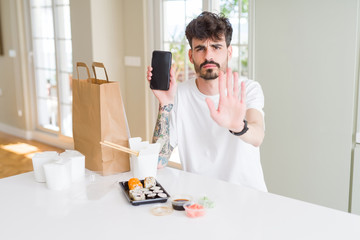 Poster - Young man eating asian sushi from home delivery and showing smartphone screen with open hand doing stop sign with serious and confident expression, defense gesture