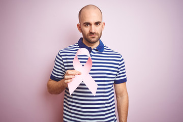 Poster - Young man holding pink brest cancer ribbon over isolated background with a confident expression on smart face thinking serious