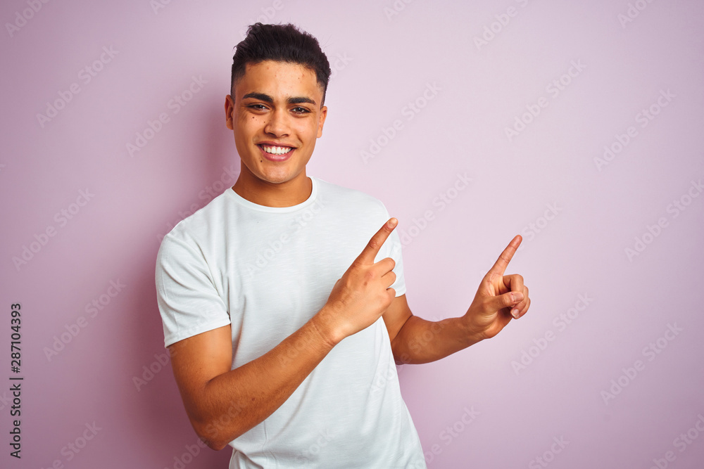 Young brazilian man wearing t-shirt standing over isolated pink background smiling and looking at the camera pointing with two hands and fingers to the side.