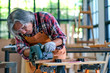 © narong - Asian elderly craftsman or carpenter use electrical saw to cut wood and generate wood dust in the room with glass windows during day light.