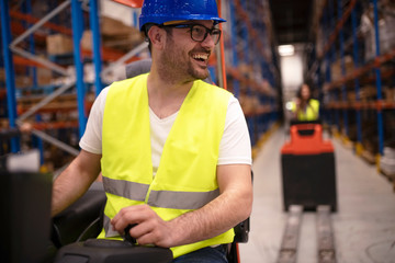 Wall Mural - Warehouse workers enjoy driving forklifts in large factory storage department. Industrial workers operating lifting machines to relocate palette with goods.