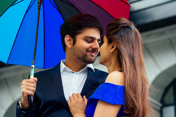 Wall Mural - indian woman in long evening blue dress spinding romantic time together with lover handsome boyfriend under multi-colored rainbow umbrella europe urban downtown city