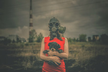Woman In Red Dress And In Gas Mask With A Toy Bear Is Standing Among Burnt Field On A Smoking Chimney Background. Air Pollution Concept.