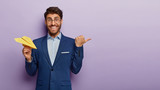 Studio shot of happy male entrepreneur wears round glasses and formal suit, holds yellow paper plane, points thumb away on blank space, demonstrates something for business partners or comapny owners