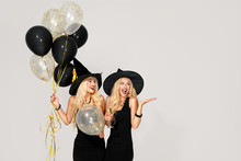 Portrait Of Two Happy Young Women In Black Witch Halloween Costumes With Balloons On Party Over White Background. The Concept Of Halloween . Funny Faces