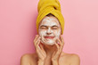Isolated shot of pleased young female touches face skin with soap, keeps eyes closed, washes her face in morning, stands naked against pink background, has spa treatment. Cleanliness concept