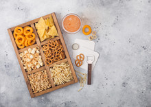 Glass Of Craft Lager Beer And Opener With Box Of Snacks On Light Background. Pretzel,salty Potato Sticks, Peanuts, Onion Rings With Nachos In Vintage Box With Openers And Beer Mats. Space For Text