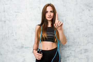 Wall Mural - Young caucasian sportswoman holding a jump rope showing number one with finger.