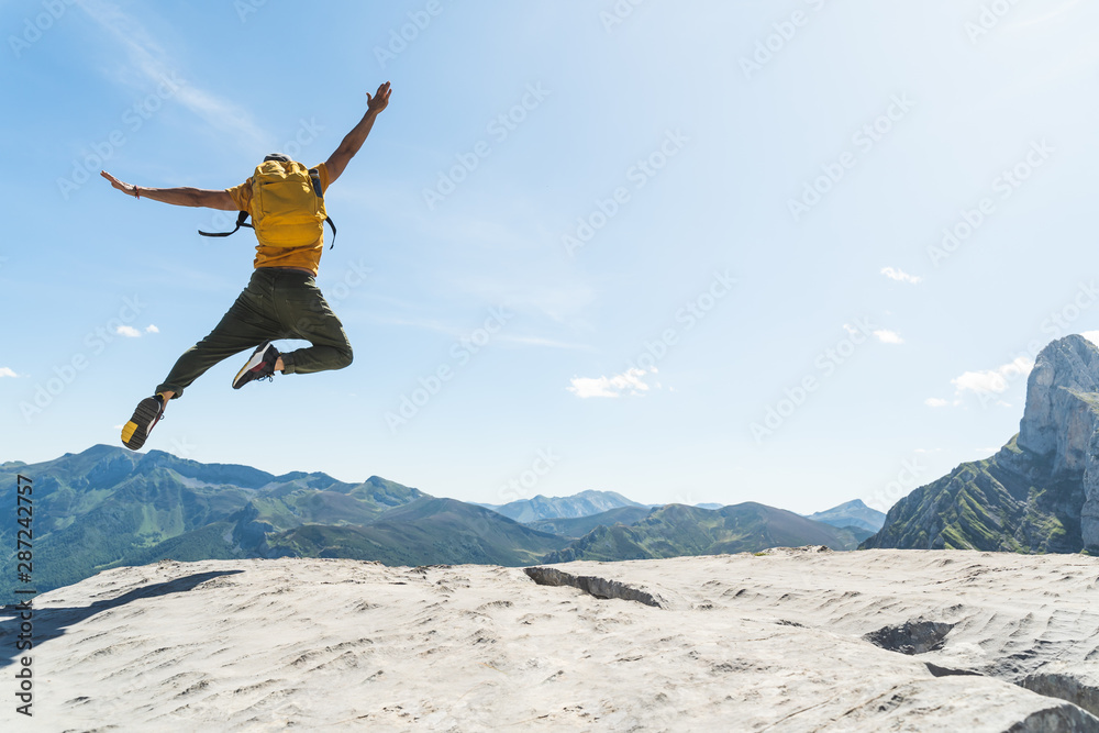 Young Man Jumping on Top of a Mountain Wearing Yellow Backpack. - obrazy, fototapety, plakaty 