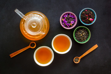 Wall Mural - Cups, teapot, dry tea on black background top view