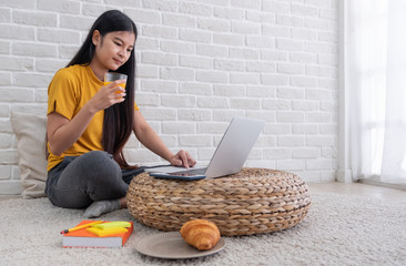Poster - Asian female sit on floor using laptop on wicker stand and drink orange juice in living room at home with relax emotion.Work at home concept.work from home.relax lifestyle