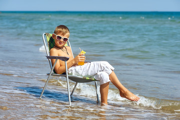 The boy is resting on the sea coast . Child sitting on a beach chair holding a glass of juice . The concept of children's recreation