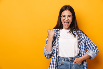 Sticker - Emotional happy young cute teenage girl in glasses posing isolated over yellow wall background showing winner gesture.