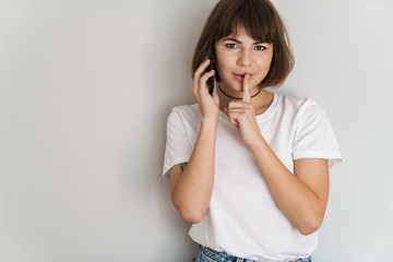 Poster - Flirty young lady isolated over grey wall background talking by mobile phone showing silence gesture.