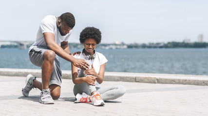 Black Guy Helping Girl Examining Sprained Ankle At Riverbank