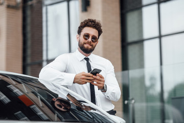 Wall Mural - Handsome, smiling, happy, bearded businessman is using his mobile phone and standing near his car outdoors on the streets. Looking to the camera.