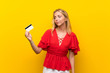 © luismolinero - Blonde young woman over isolated yellow background holding a credit card