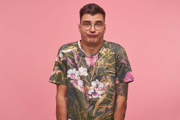 Canvas Print - Indoor shot of puzzled young male in casual t-shirt posing over pink background with hands down, making perplex face, looking to camera with pursed lips