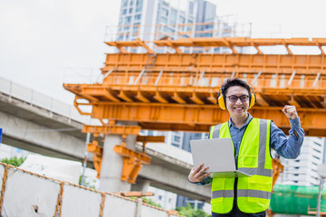 Wall Mural - Image outside the industrial construction engineers in yellow protective ear muff discuss new project while using laptop and happy smile on the open building site near the crane.