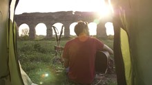 Young Man Backpacker Playing Guitar Outside Camping Tent Sitting On Grass In Front Of Roman Aqueduct Arches In Parco Degli Acquedotti Park Ruins In Rome At Sunset Sticks In Ground Camera Shot Inside