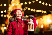 Holidays, Childhood And People Concept - Happy Little Girl With Lantern At Christmas Market In Winter Evening