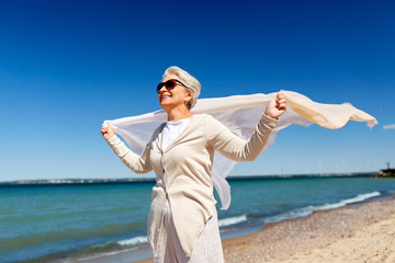 Wall Mural - people and leisure concept - portrait of happy senior woman in sunglasses with waving scarf on beach in estonia