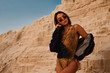 Young beautiful woman with luxury tanned body wearing snakeskin print swimwear, denim jacket, trendy sunglasses, ethnic necklace, posing in sand desert, at sunset. Copy, empty space for text