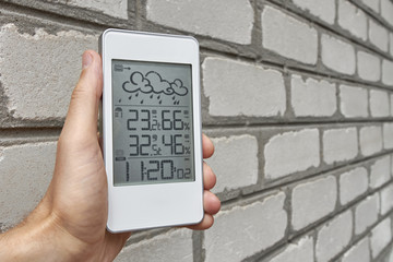 Wall Mural - Best personal weather station device with weather conditions inside and outside. A man holds a gadget in his hand against a brick wall.