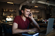 © ZoFot - Smiling business man talking with client via mobile phone and reading information on laptop computer while sitting in coffee shop during work break. Happy hipster guy having smartphone conversation