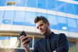 © ZoFot - Young successful man online ordering via cellphone while standing outside company during work break. Bearded male entrepreneur reading payment notification on mobile phone, standing near enterprise