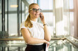 © ZoFot - Smiling woman in official wear successful government worker talking via smartphone while standing in modern interior during break at job. Joyful female in glasses skilled entrepreneur phoning