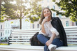 © shurkin_son - Low angle view of positive stylish middle aged female in trendy leather jacket, blue jeans and sneakers having pensive look, sitting comfortably on bench with cell phone, thinking where to have lunch