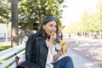 Wall Mural - Food, travel, people and lifestyle concept. Side view of confident stylish middle aged female enjoying happy peaceful days of her retirement, sitting comfortably on bench, eating ice cream