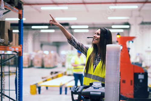 Cuadros en Lienzo Female forklift operator with headset communication equipment showing location on the shelf to place packages in large warehouse storage center while worker organizing distribution in background