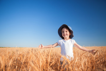 Wall Mural - Happy girl walking in golden wheat, enjoying the life in the field. Nature beauty, blue sky and field of wheat. Family outdoor lifestyle. Freedom concept. Cute little girl in summer field