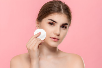 Wall Mural - Beautiful model applyes cosmetic product to skin with white cotton sponge. Model looks at the camera isolated over pink background. Concept of beauty and health treatment.