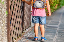 Rear View Of A 3-4 Year Old Child Standing At Wooden Rustic Fence With A Rucksack On The Sidewalk On A Sunny Summer Day