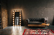 Corner room with a wooden and  gray cement wall, an aged floor, black glass doors and electric dressing makeup mirror, lights bulbs tern on, sofa. Loft style