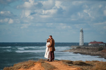 Romantic Holiday Concept Of Two Young People In Autumn Clothes Hugging Each Other On Scenic Landscape With Stormy Ocean With Lighthouse On Cloudy Background