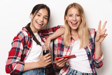 Wall Mural - Cheerful happy positive pleased young pretty girls friends sisters posing isolated over white wall background using mobile phone.