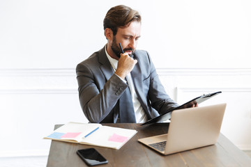 Canvas Print - Image of thinking handsome businessman reading documents while working at laptop in office