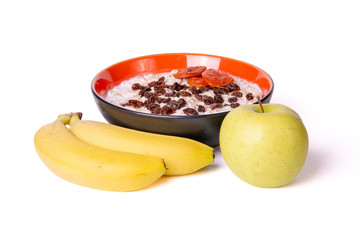 Sprouted wheat porridge sprinkled with raisins and dried apricots in red and black bowl with green apple and yellow bananas near it. Isolated on white background. Idea for healthy diet breakfast.