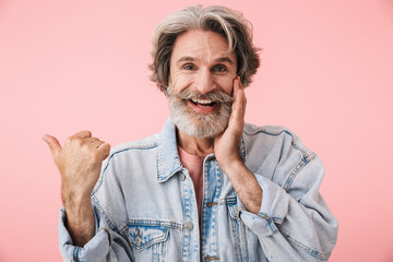 Portrait of joyful old man with gray beard laughing and pointing finger at copyspace