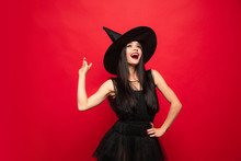 Young Brunette Woman In Black Hat And Costume On Red Background. Attractive Caucasian Female Model. Halloween, Black Friday, Cyber Monday, Sales, Autumn Concept. Copyspace. Pointing Up.