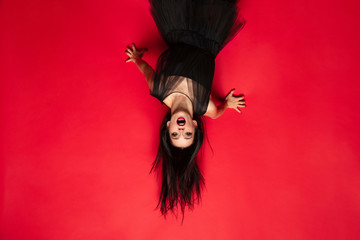 Wall Mural - Young brunette woman in black hat and costume on red background. Attractive caucasian female model. Halloween, black friday, cyber monday, sales, autumn concept. Scary screaming upside down.