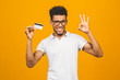 © denis_vermenko - Afro american customer man holding credit card standing over isolated yellow background happy with big smile doing ok sign, thumb up with fingers, excellent sign
